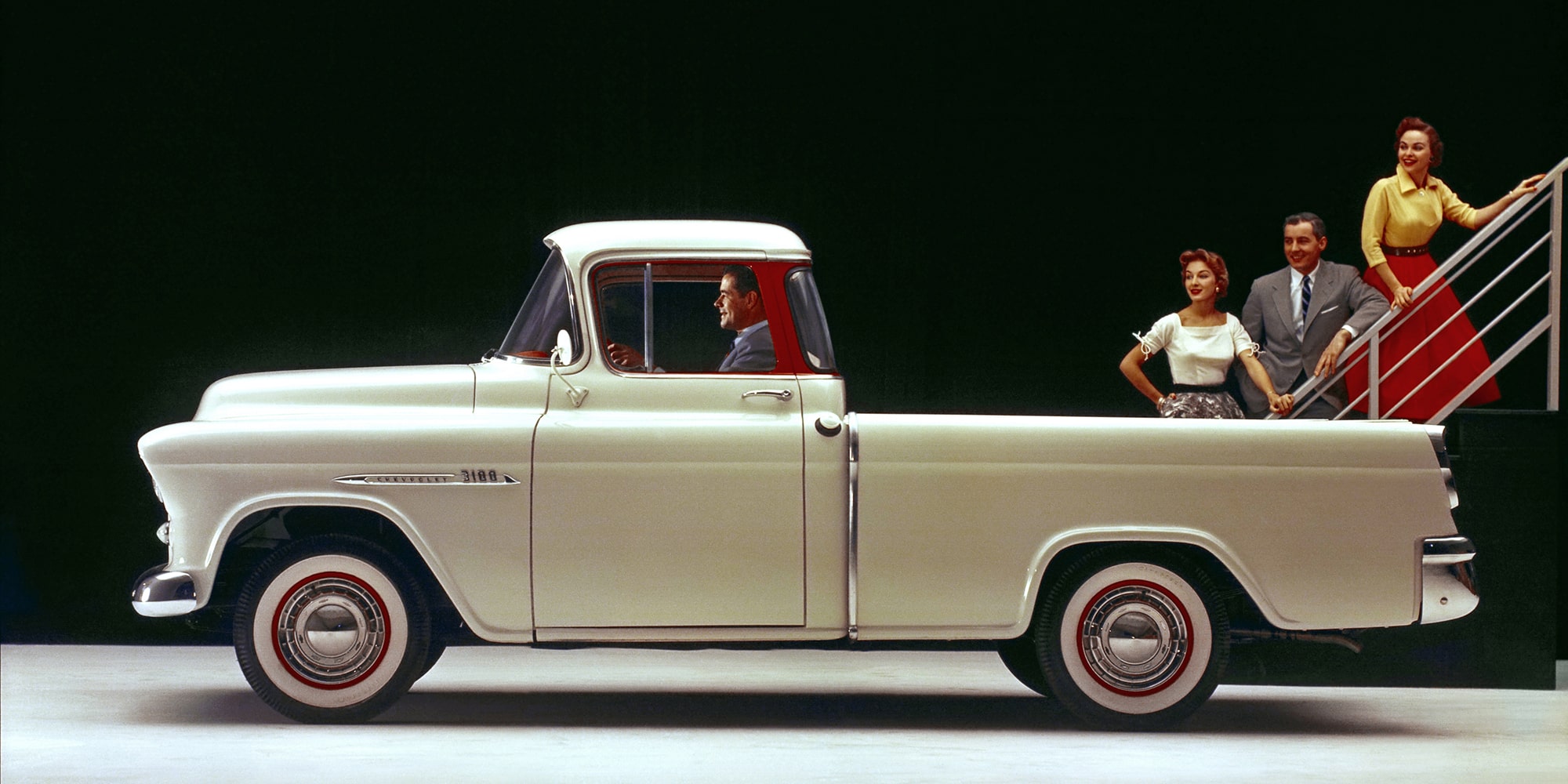 Vista de perfil de una pickup clásica Chevrolet Cameo Carrier 1955 en Marfil con detalles en Rojo y neumáticos de banda blanca
