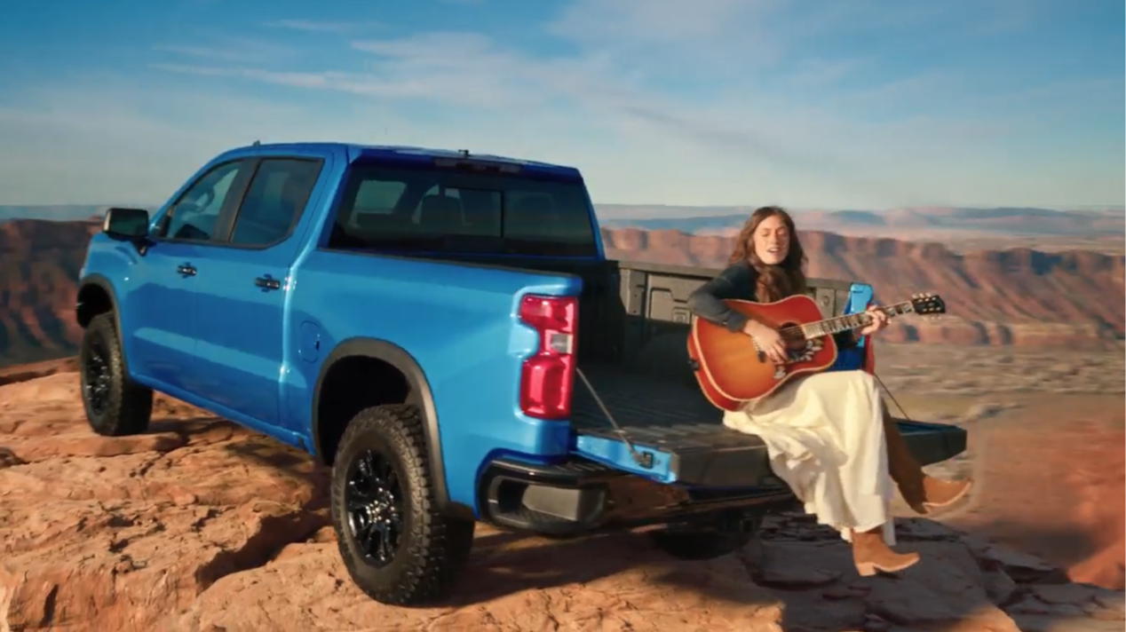 Una mujer cantando y tocando la guitarra sentada en la caja de una camioneta Chevy Azul estacionada sobre un acantilado en las montañas