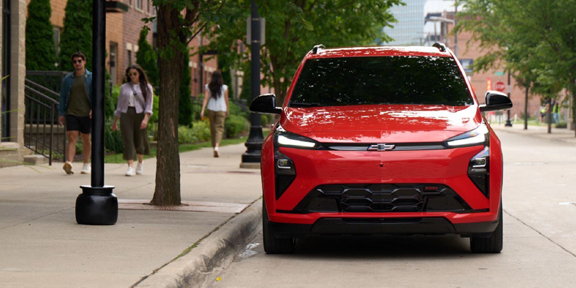 Vista delantera de una Chevy Bolt Roja estacionada en una calle llena de árboles con peatones caminando a su lado