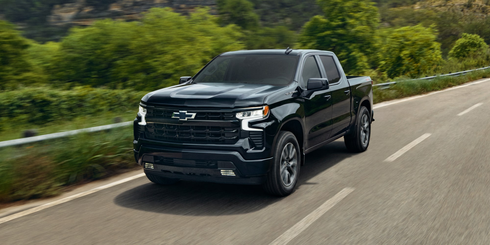 Vista delantera de tres cuartos de una Chevy Silverado 1500 2026 Negro circulando por un camino pavimentado lleno de follaje verde