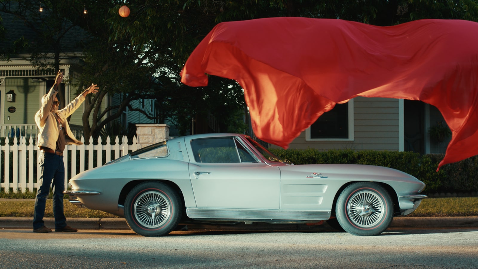 A Classic Corvette Sting Ray Revealed and Parked on a Suburban Street While a Man Throws Off a Red Car Cover