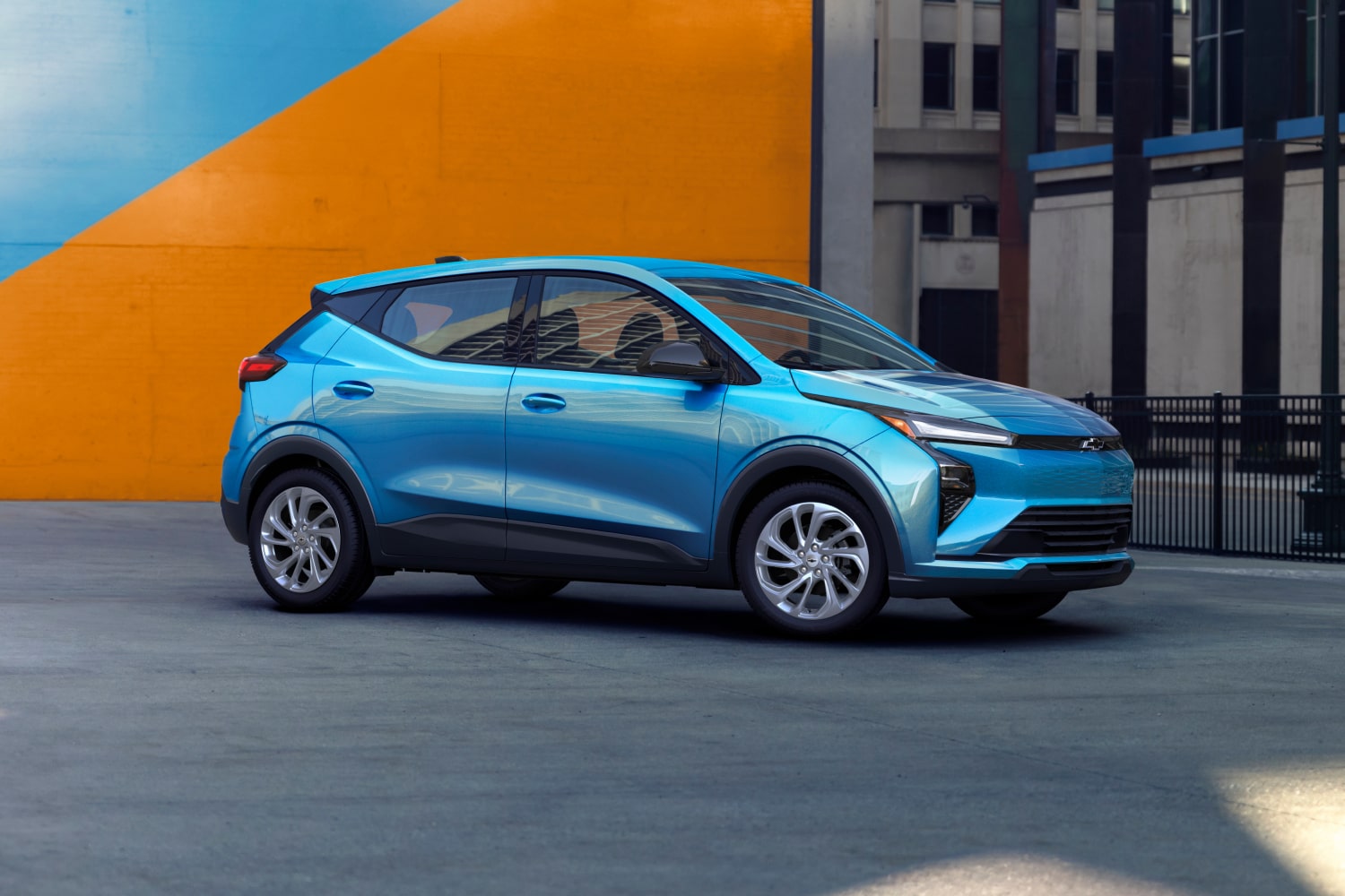 Una Chevrolet Bolt azul estacionada contra una pared geométrica colorida en un entorno urbano.