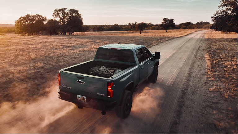 A Chevy Silverado Pickup Truck Driving Along a Rural Gravel Road Through Open Countryside With Dust Trailing Behind the Vehicle at Sunset.