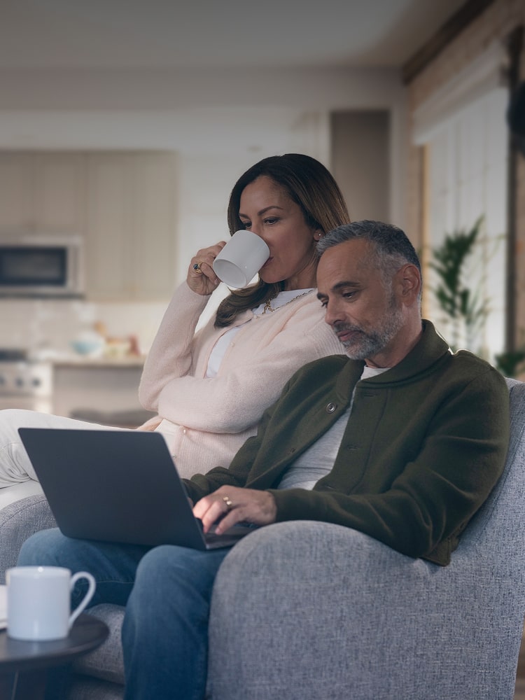 Two people at a table using a laptop in a bright room with large windows overlooking trees and a lawn. Text on the image reads: “BUY YOUR WAY. Your Chevy, on your time and terms.”