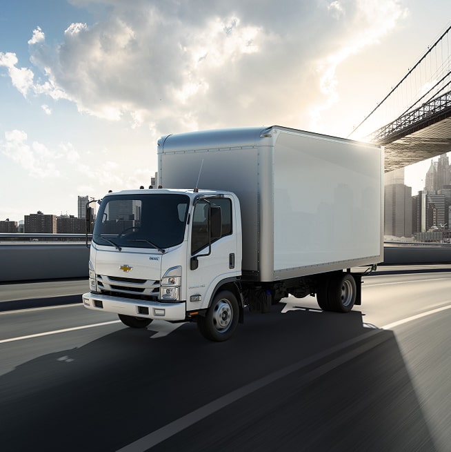 Front Angled View of a 2026 Chevy Low Cab Forward Commercial Truck Driving Down a Highway with a City in the Background.