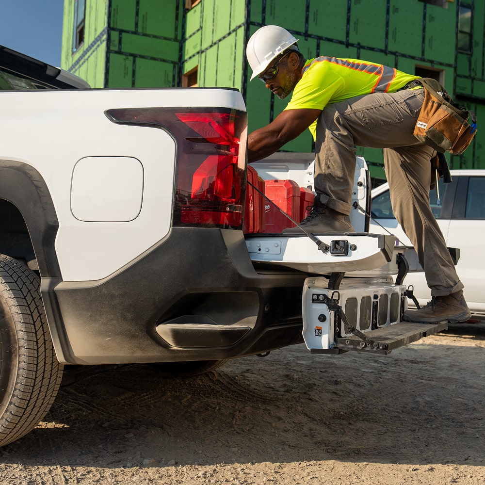 A Construction Worker Accessing Equipment From the Multi-Flex Midgate and Open Tailgate of a White Commercial Chevy Silverado EV.