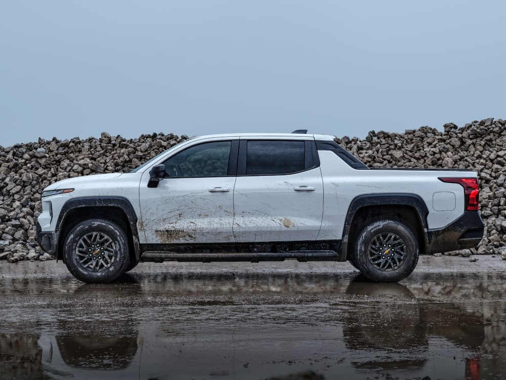 A White Commercial Chevy Silverado EV Parked on a Wet Road With Rocks Piled in the Background.