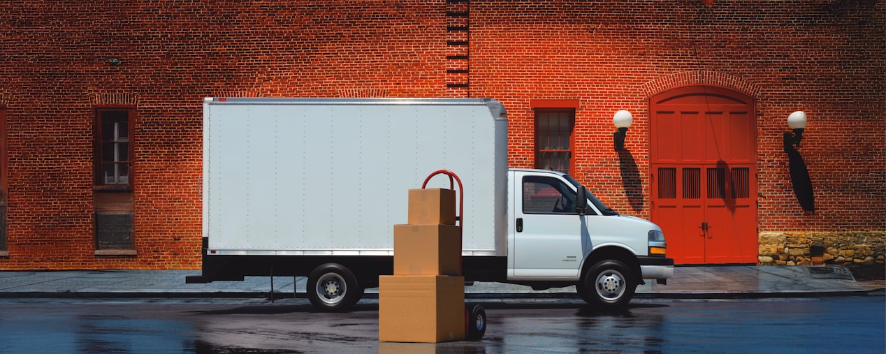 A 2026 Chevrolet Express Cutaway Box Truck Parked on a Wet Street in front of a Brick Building with Stacked Boxes.