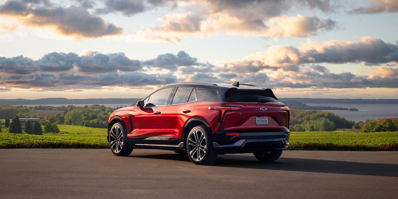 A Red Chevrolet Blazer EV Driving on a Scenic Road With Rolling Green Hills and a Dramatic Cloud-Filled Sky in the Background.