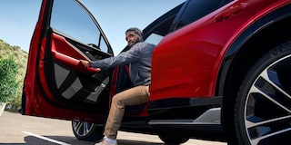 A Person Entering a Red Chevrolet Blazer EV Through the Driver’s Side Door With the Vehicle Parked on a Sunlit Road and Hills in the Background.