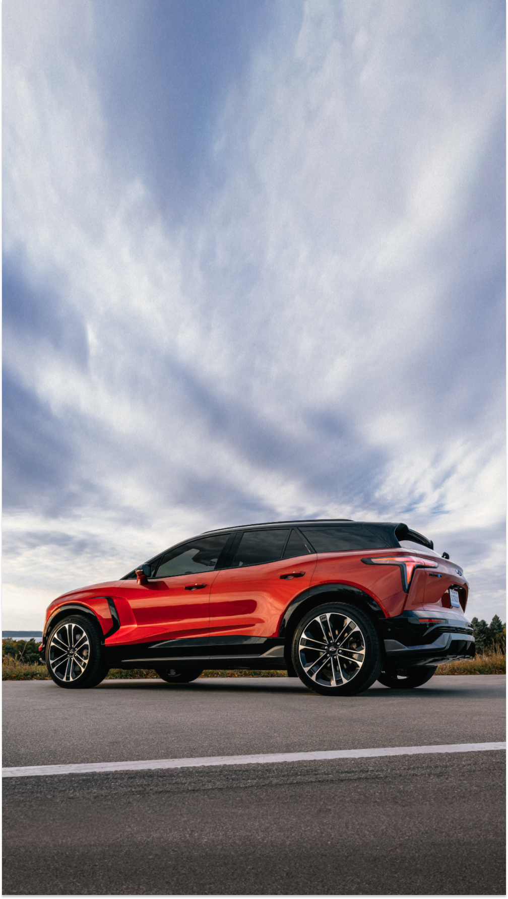 A Red Chevrolet Blazer EV Driving on an Open Road With a Vast Blue Sky and Wispy Clouds Above and Trees in the Distance.