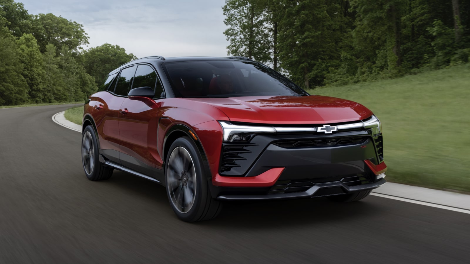 A Red Chevrolet Blazer EV Driving on a Tree-Lined Road With Green Grass and Foliage in the Background.