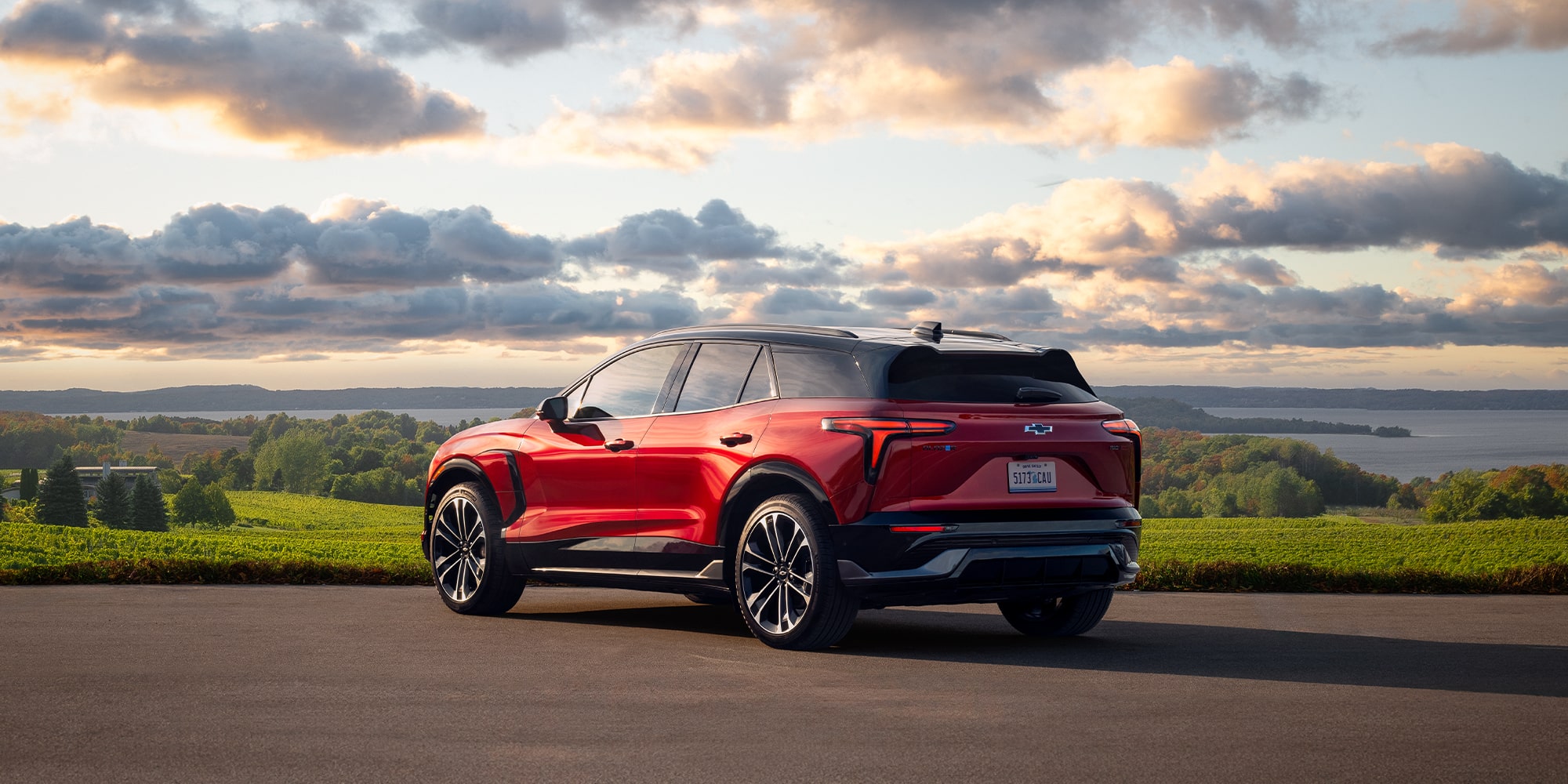A Red Chevrolet Blazer EV Driving on a Scenic Road With Rolling Green Hills and a Dramatic Cloud-Filled Sky in the Background.