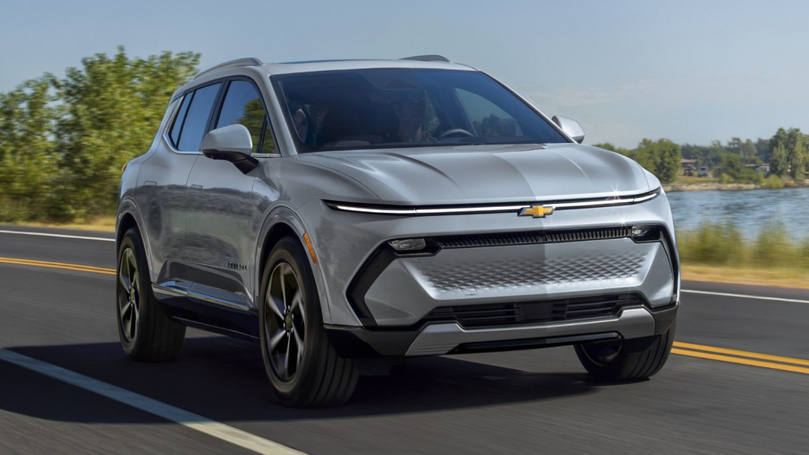 A Silver Chevrolet Equinox EV Driving on a Road Next to a Body of Water and Trees on a Sunny Day.