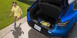 A Blue Chevrolet Equinox EV With Its Trunk Open, Revealing a Child in a Yellow Shirt Playing Nearby on a Driveway.