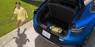 A Blue Chevrolet Equinox EV With Its Trunk Open, Revealing a Child in a Yellow Shirt Playing Nearby on a Driveway.