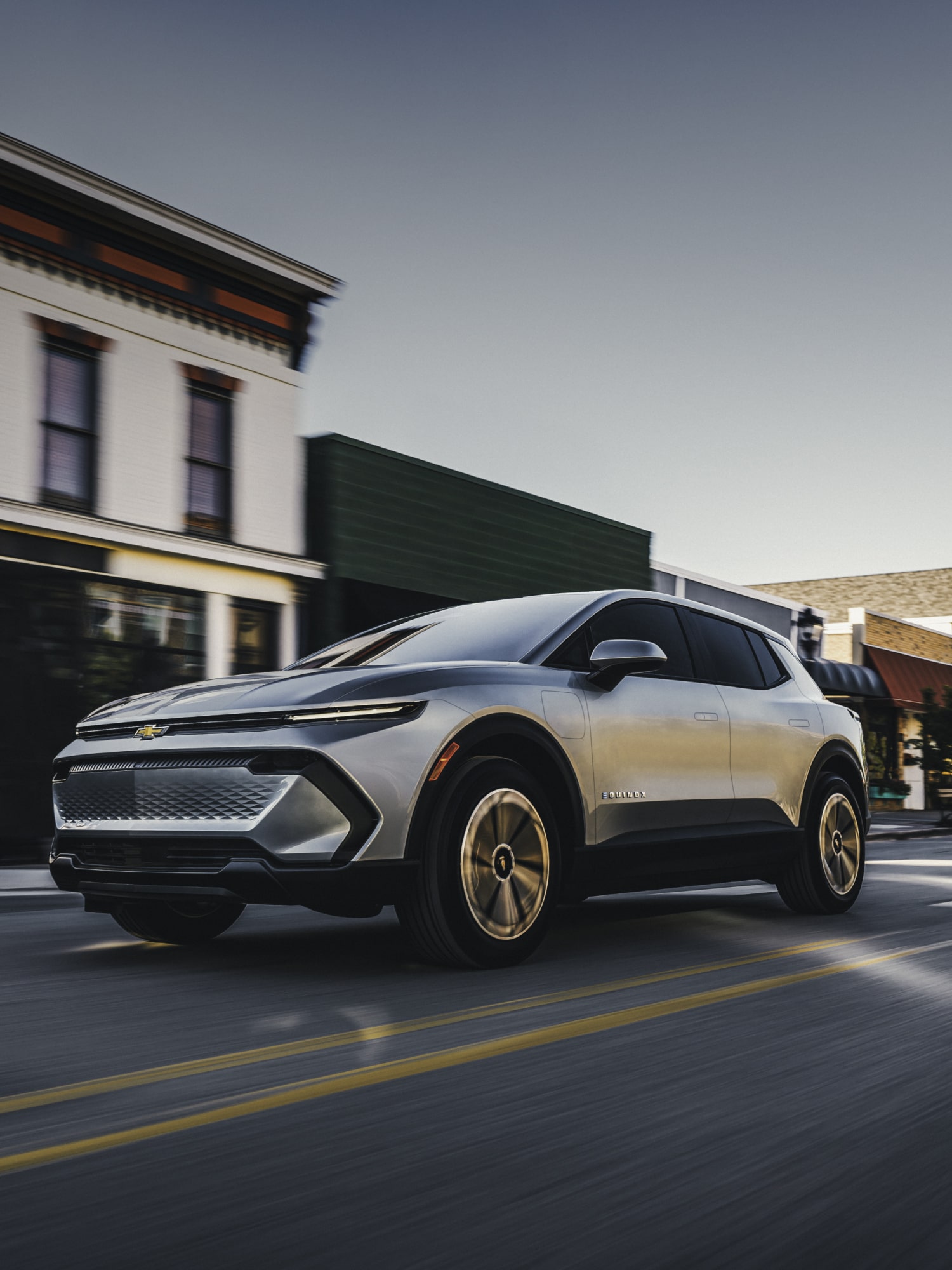 A Silver Chevrolet Equinox EV Driving Through a Quaint Town Street Lined With Historic Buildings.