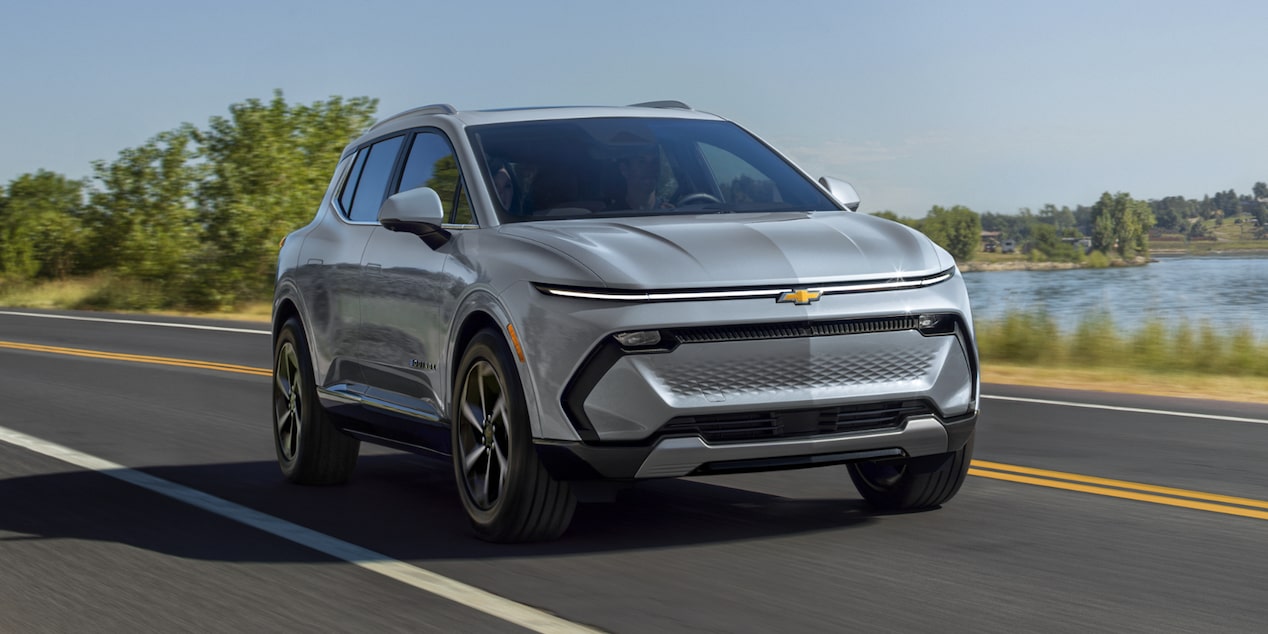 A Silver Chevrolet Equinox EV Driving on a Road Next to a Lake and Green Trees.