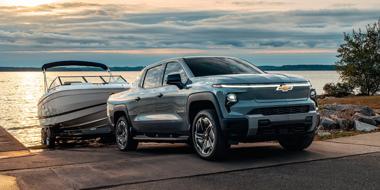 Vista de medio perfil de una Chevy Silverado EV 2026 retirando una lancha de la orilla de un cuerpo de agua, por la tarde