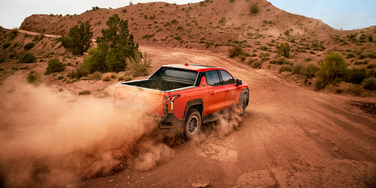 A Habanero Orange Chevrolet Silverado EV Trail Boss Pickup Truck Drives On A Dirt Road, Kicking Up A Large Cloud Of Dust Behind It.