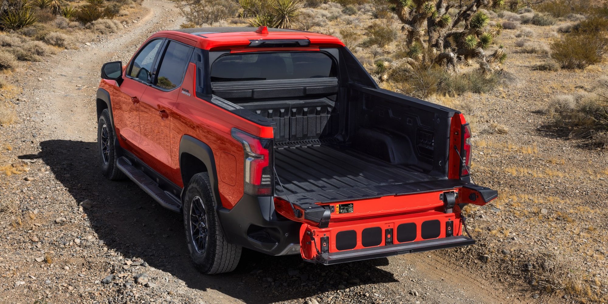 Three Quarters Rear View of an Orange 2026 Chevy Silverado on a Rocky Road under a Sunny Day