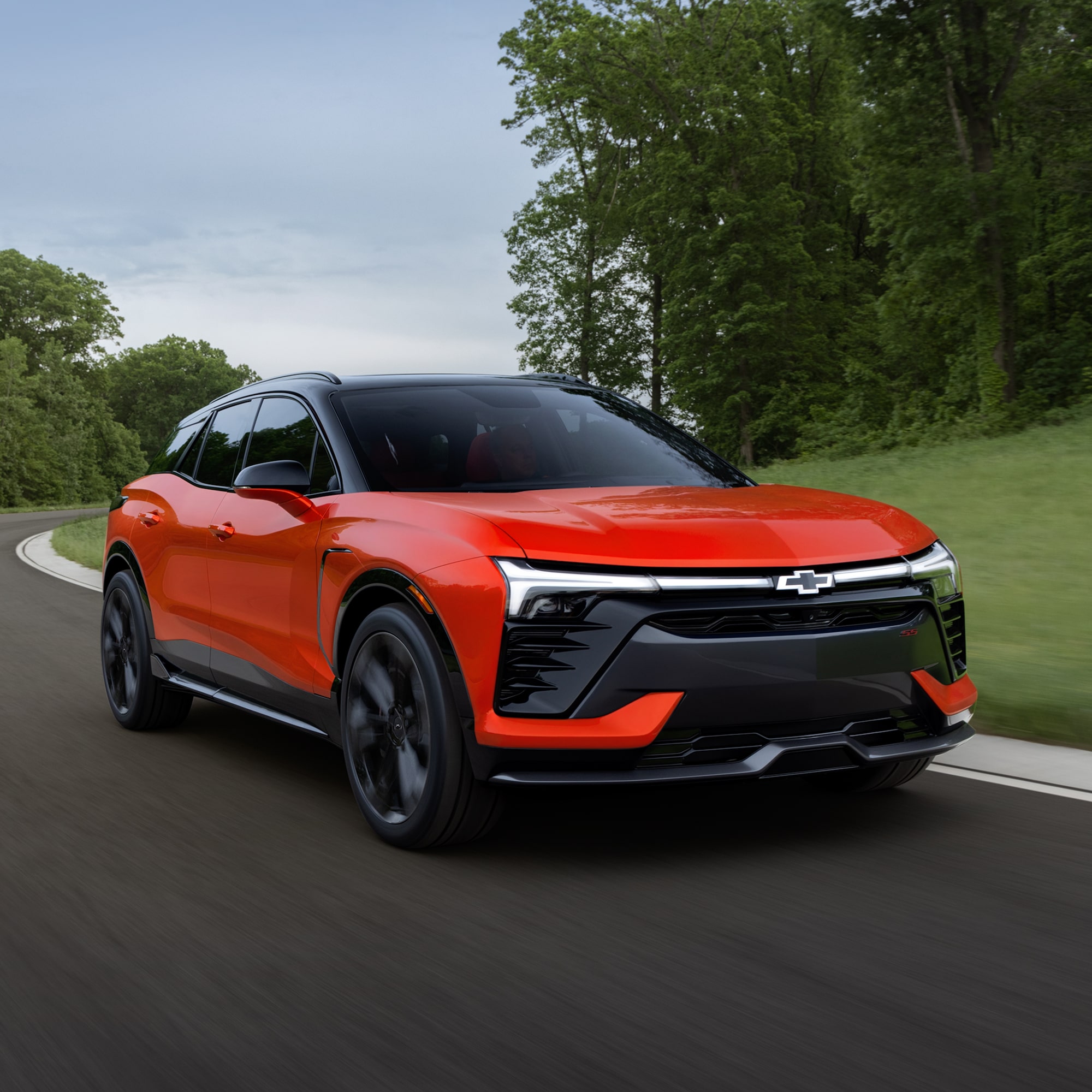 A 2027 Chevy Blazer EV SS Driving Along a Tree-Lined Road Showcasing Distinctive LED Lighting, Black Alloy Wheels, a Contrasting Roof, and Athletic Exterior Styling From a Front Three-Quarter Angle.