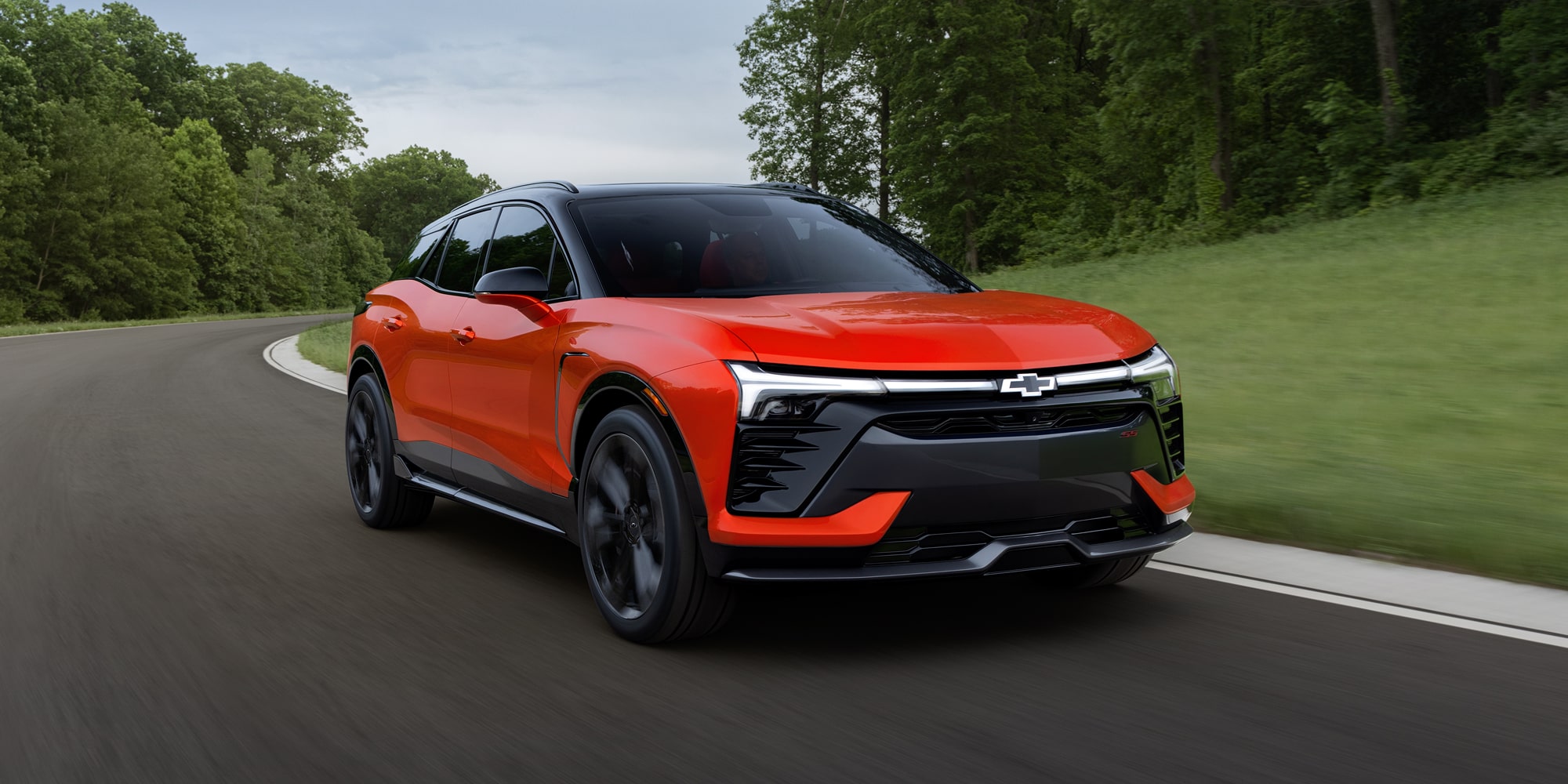 A 2027 Chevy Blazer EV SS Driving Along a Tree-Lined Road Showcasing Distinctive LED Lighting, Black Alloy Wheels, a Contrasting Roof, and Athletic Exterior Styling From a Front Three-Quarter Angle.
