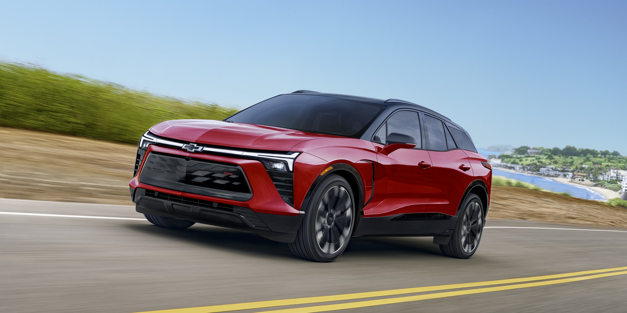 A 2027 Chevy Blazer EV RS Driving Along a Scenic Road With Black Alloy Wheels, a Contrasting Roof, and Distinctive Front-End Styling Visible From a Front Three-Quarter Angle.