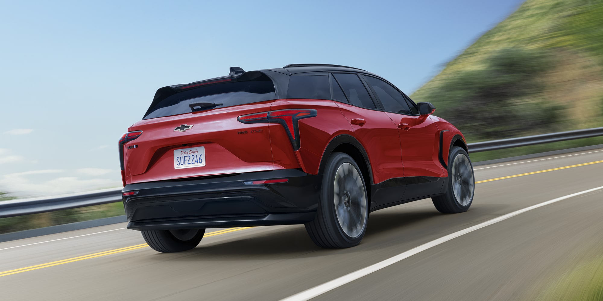 A 2027 Chevy Blazer EV RS Driving Along a Winding Road With LED Taillights, a Rear Roof Spoiler, and Sculpted Exterior Styling Visible From a Rear Three-Quarter Angle.