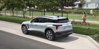 A 2027 Chevy Blazer EV LT Driving Along a Residential Street With Aerodynamic Styling, LED Taillights, and a Modern Exterior Design Visible From a Rear Three-Quarter Angle.