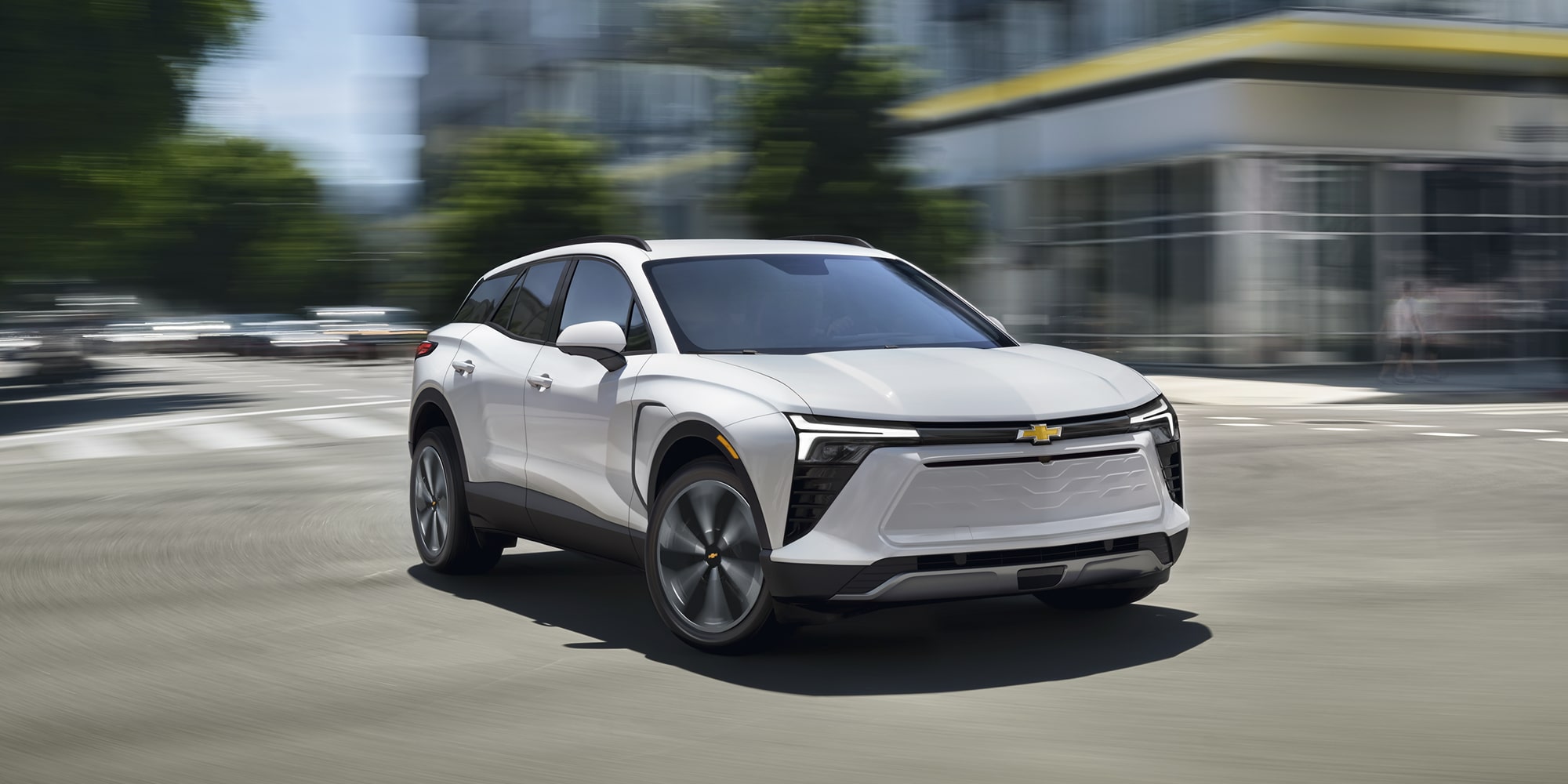 A 2027 Chevy Blazer EV LT Driving Through an Urban Environment With Distinctive LED Lighting, Black Alloy Wheels, and a Streamlined Exterior Design Visible From a Front Three-Quarter Angle.