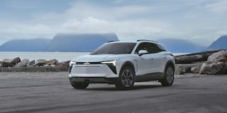 A 2027 Chevy Blazer EV LT Parked on a Coastal Shoreline With Black Alloy Wheels, a Contrasting Roof, and Modern Exterior Styling Visible From a Front Three-Quarter Angle Against a Mountainous Waterfront Backdrop.