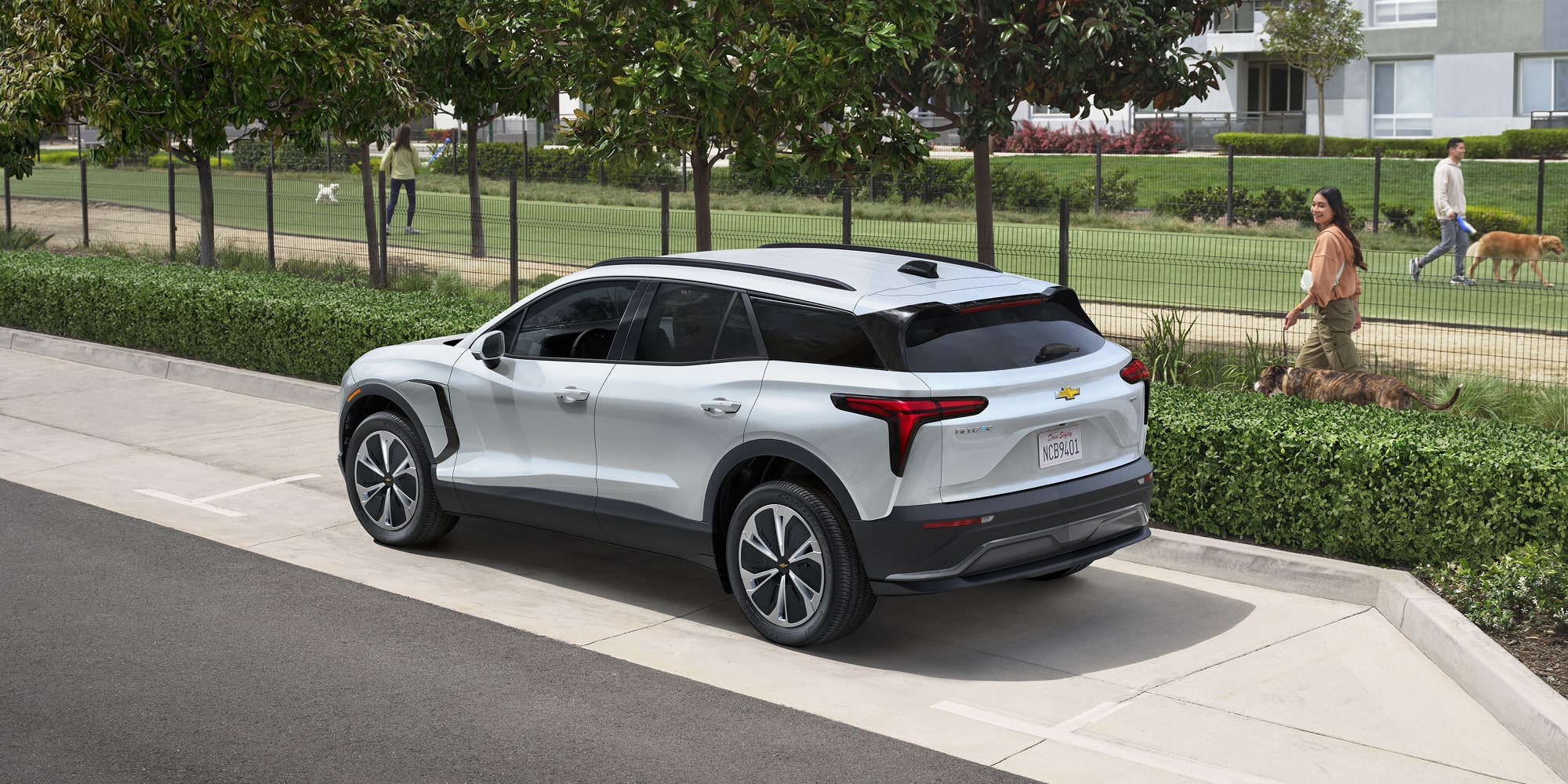 A 2027 Chevy Blazer EV Driving Along a Residential Street With Sleek Body Lines, Aerodynamic Styling, and LED Taillights Visible From a Rear Three-Quarter Angle.