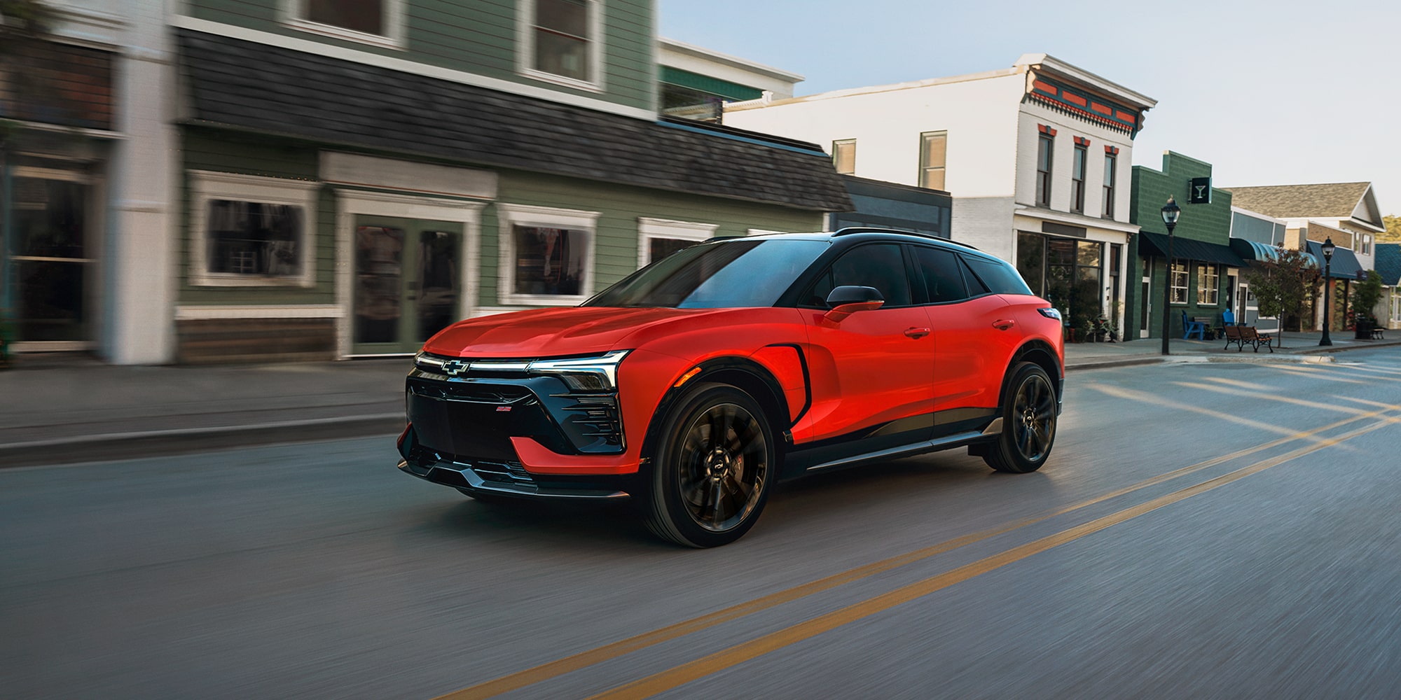 A 2027 Chevy Blazer EV Driving Through a Downtown Street With Black Alloy Wheels, a Contrasting Black Roof, and Sporty Front-End Styling Visible From a Front Three-Quarter Angle.