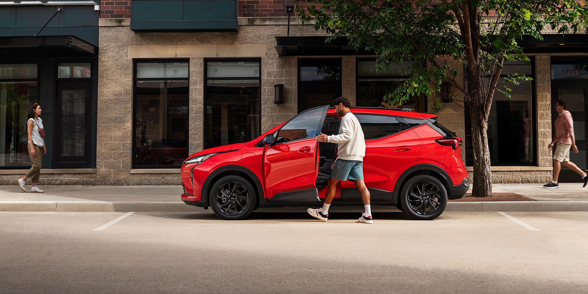 The Side View of a Red 2027 Chevrolet Bolt, with a Man Getting into the Driver's Side Door on a City Street.