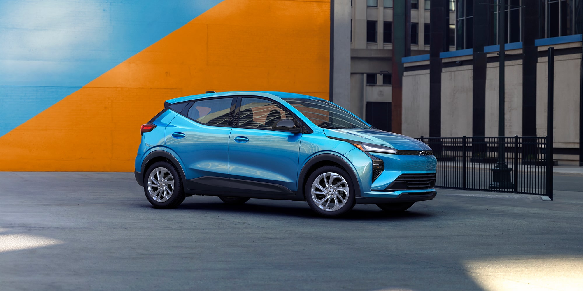 A Light Blue 2027 Chevrolet Bolt is Parked in a City Setting in Front of a Large Orange and Blue Wall.