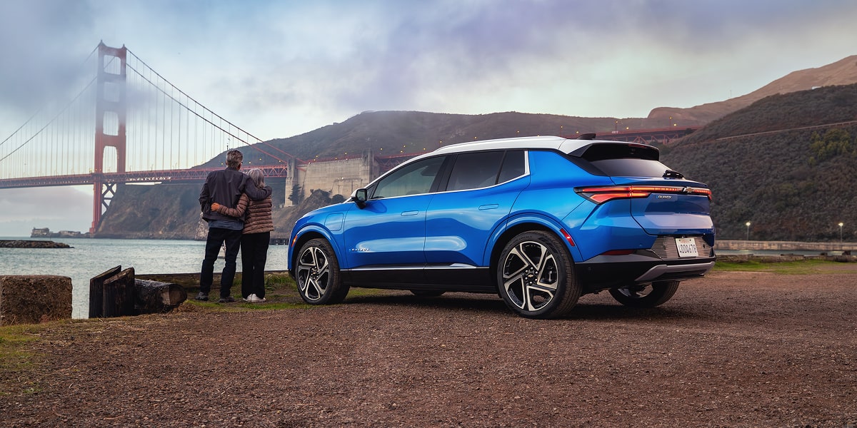 Vista trasera de perfil de Equinox EV Azul estacionada mientras una pareja mira hacia el puente Golden Gate