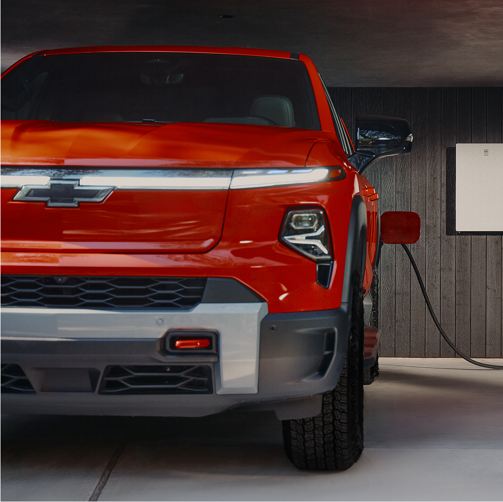 A Close-Up of the Front End of a Red Chevy Silverado EV Parked Beside a Home Charging Station.