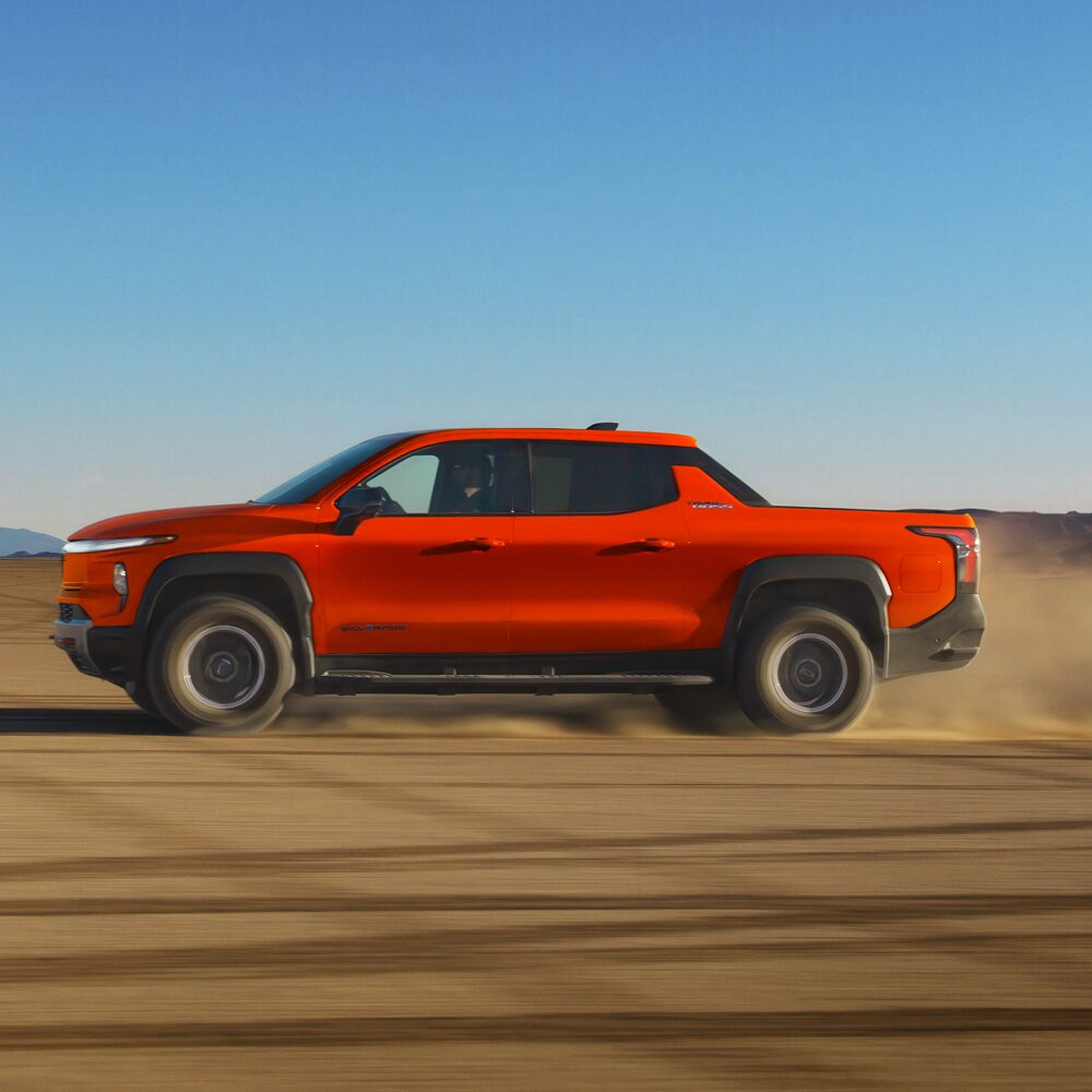 A Red Chevy Silverado EV Driving Across a Sandy Off-Road Trail With Dust Kicking Up Behind the Vehicle.