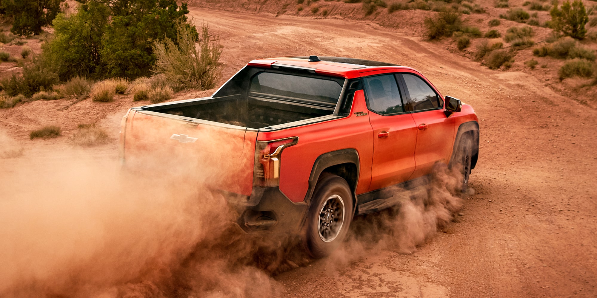 A Habanero Orange Chevrolet Silverado EV Trail Boss Pickup Truck Drives On A Dirt Road, Kicking Up A Large Cloud Of Dust Behind It.