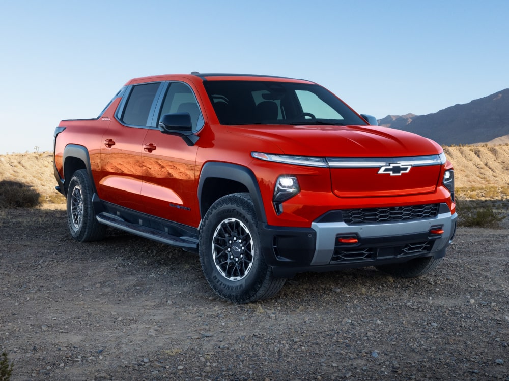 A Red Chevy Silverado EV Parked on a Dirt Road With Mountains Visible in the Background.