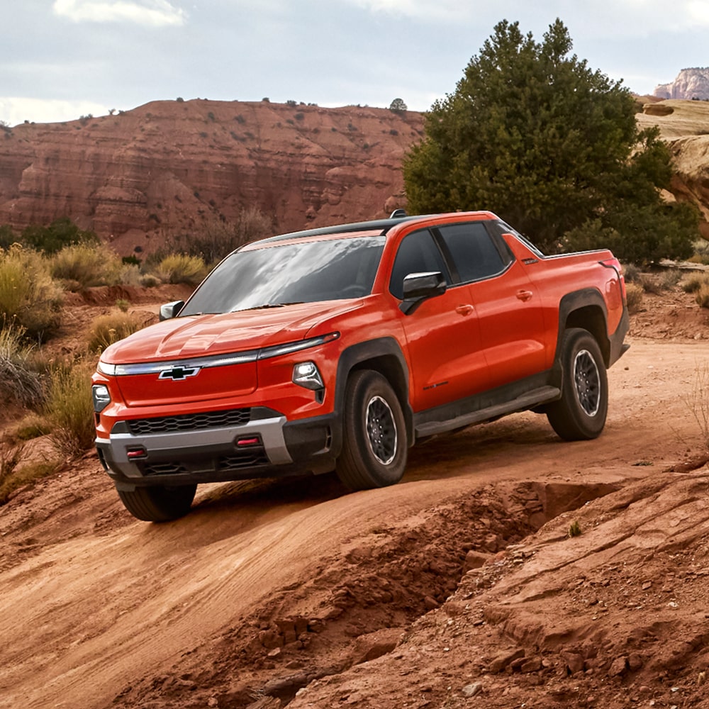 A Red Chevy Silverado EV Driving Along a Rugged Dirt Trail With Hills in the Background.