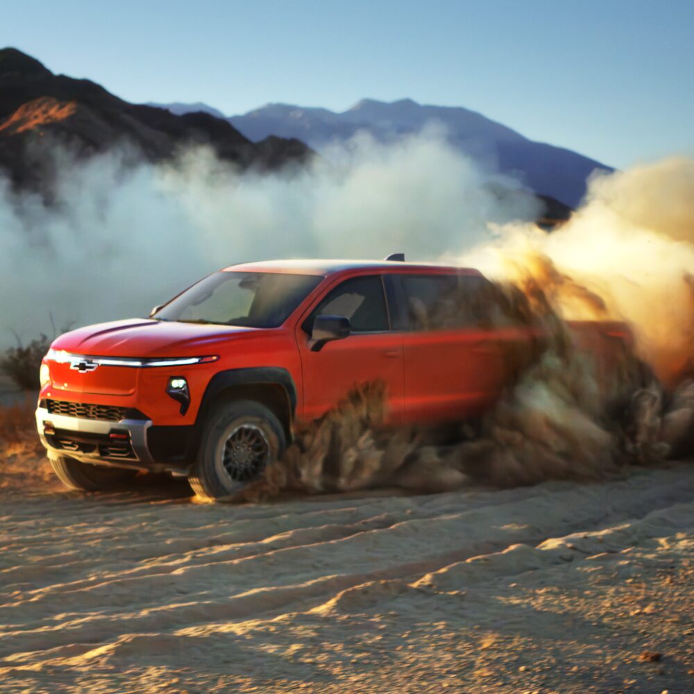 A Red Chevy Silverado EV Driving Through a Rugged Off-Road Landscape With Dust and Rocks Surrounding the Vehicle.