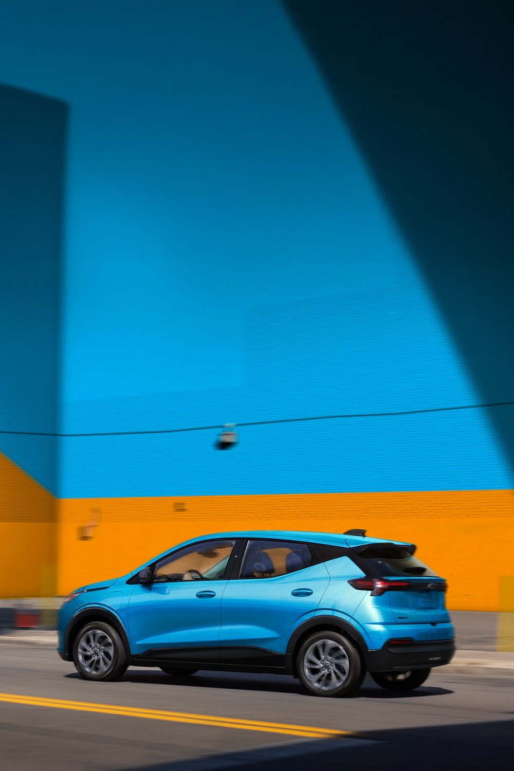 Blue 2027 Chevrolet Bolt driving aside an orange and blue  wall