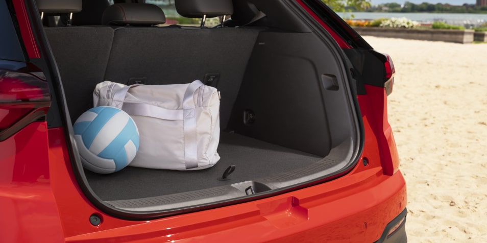 The Trunk of a Red 2027 Chevrolet Bolt is Open on a Sandy Beach, with a Blue and White Volleyball and a White Duffel Bag Inside