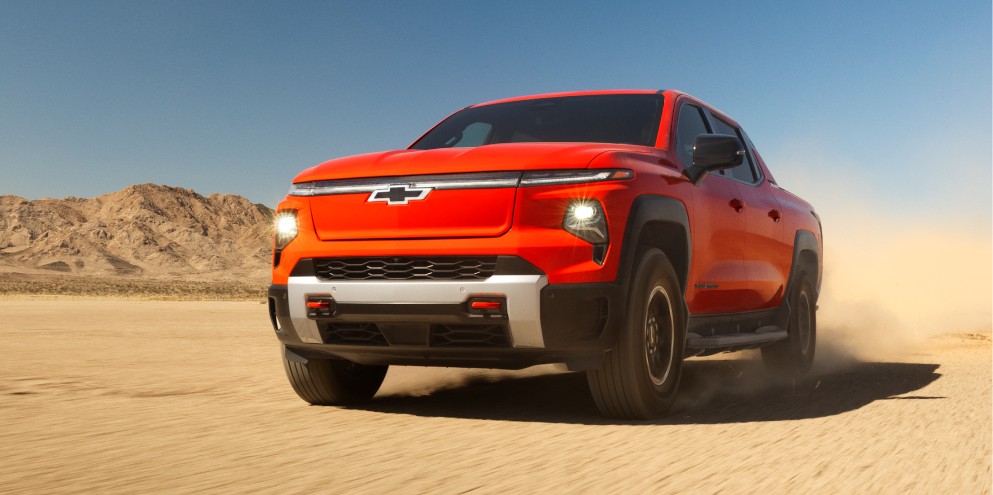 Front Quarter View of an Orange Chevrolet Silverado EV Driving Forward on a Dirt Road With Headlights on.