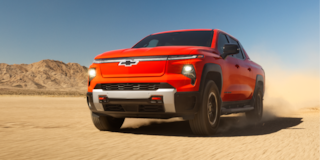 Front Quarter View of an Orange Chevrolet Silverado EV Driving Forward on a Dirt Road With Headlights on.