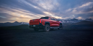 Rear View of an Orange Chevrolet Silverado EV Parked on Rocky Terrain With Mountains in the Background.