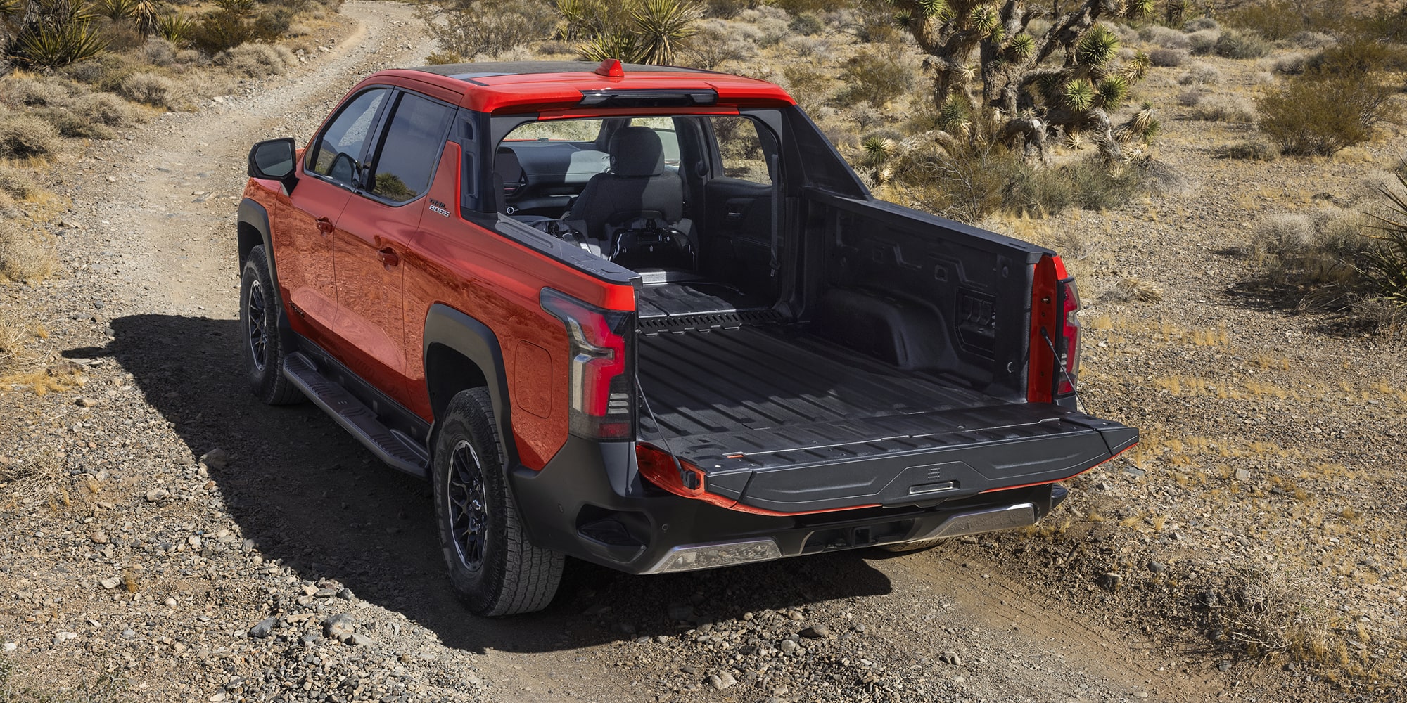 Rear View of an Orange Chevrolet Silverado EV Parked on a Dirt Trail With the Multi-Flex Midgate Open, Showing the Empty Bed.