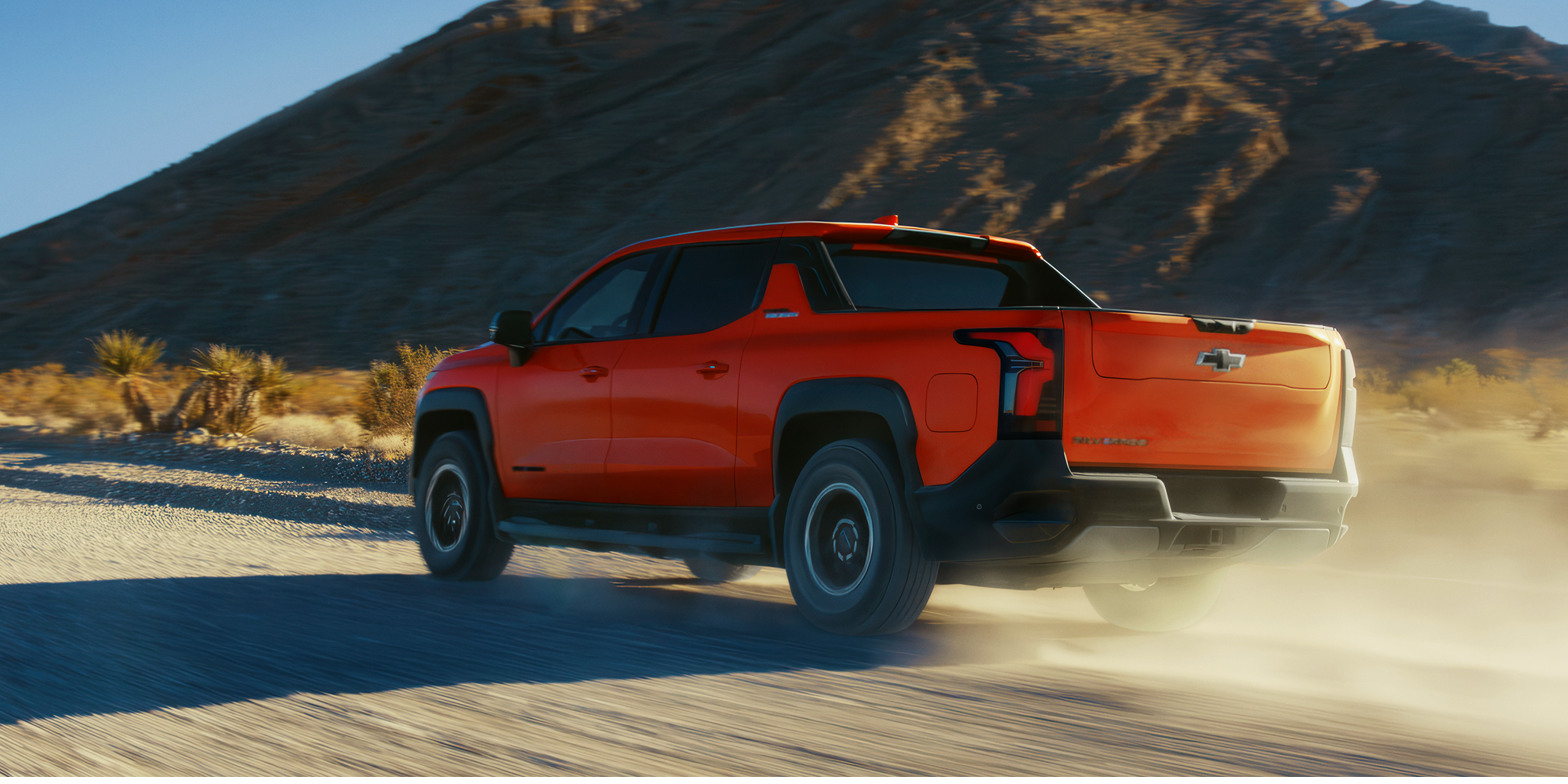 Rear Quarter View of an Orange Chevrolet Silverado EV Driving on a Dirt Road, Trailing Dust in a Mountainous Desert.